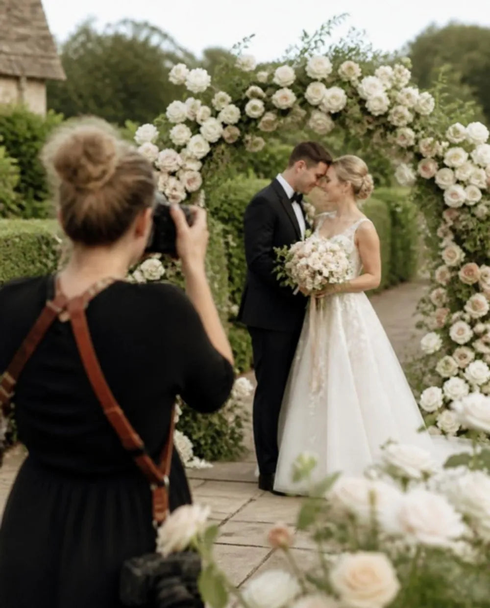 A photographer taking a picture of a bride and groom under an arch of roses
